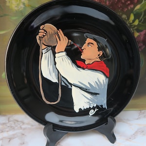 Peut inclure: Assiette décorative noire avec une image peinte à la main d'une personne en chemise blanche, écharpe rouge et béret, buvant dans une poche en cuir. L'assiette est présentée sur un présentoir noir.