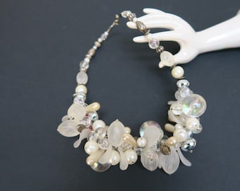 May include: A white and silver beaded necklace with a cluster of white and iridescent beads in the center. The necklace is displayed on a white hand.