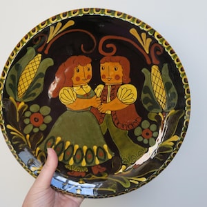 May include: A round ceramic plate with a dark brown background and a colorful folk art design. The design features two figures in traditional clothing, corn stalks, and floral motifs. The plate has a gold rim.