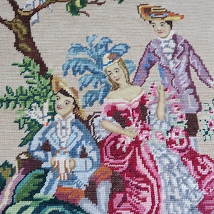 May include: A vintage needlepoint wall hanging depicting three figures in a garden setting. The figures are dressed in 18th-century attire and are surrounded by flowers and foliage. The central woman is wearing a pink and red dress, while the two men are wearing blue and purple suits. The needlepoint is worked in a variety of colours and textures, creating a realistic and detailed scene.
