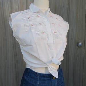 May include: White short-sleeved button-up shirt with a Peter Pan collar and pink floral embroidery. The shirt is tied at the waist.