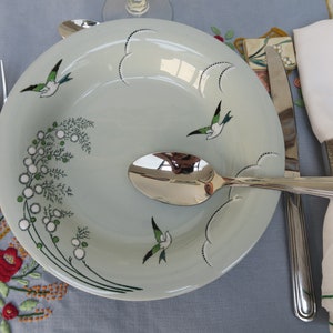 May include: A light blue ceramic dinner plate with a green and white floral design and a bird motif. The plate is set on a blue tablecloth with a silver fork, spoon, and knife. A white napkin with a silver napkin ring is on the right side of the plate.