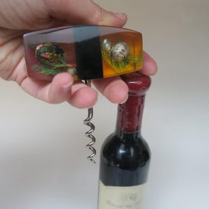 Puede incluir: Un abridor de botellas de vino decorativo con un mango rectangular con obras de arte incrustadas. El mango es una mezcla de ámbar y negro, con un sacacorchos plateado. Una botella de vino tinto está en el fondo.
