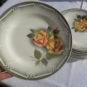May include: A set of six white ceramic plates with a green and white border. The plates feature a floral design with yellow and orange roses.