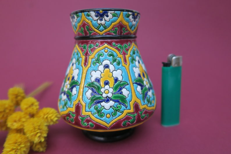 May include: A small, colorful ceramic vase with a geometric and floral design. The vase is decorated with blue, green, yellow, white, and red paint.