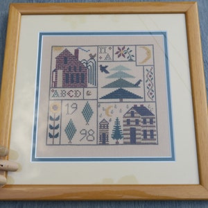 Peut inclure: Un sampler de point de croix encadré avec un fond beige représentant une maison, un arbre, un tournesol et d'autres motifs. Le sampler comprend les chiffres 19 et 98, ainsi que les lettres ABCD.