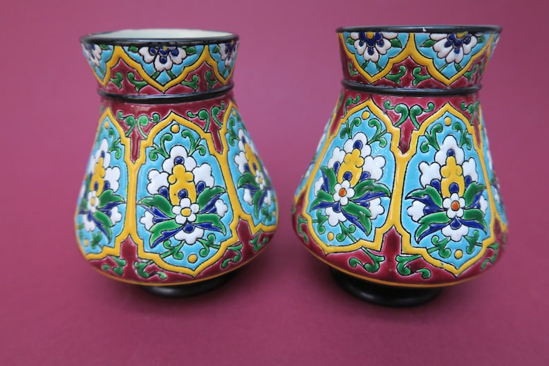 May include: Two ceramic vases with a colorful floral pattern. The vases are decorated with blue, green, yellow, white, and red flowers on a blue background. The vases have a black base.