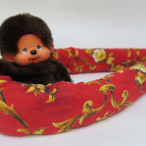 May include: A brown plush monkey toy with a red and gold floral patterned pillow. The monkey is sitting in the pillow, which is shaped like a horseshoe.
