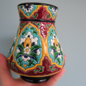 May include: A colorful ceramic vase with a geometric pattern of blue, green, yellow, white, and red flowers. The vase has a wide base and a narrow neck.