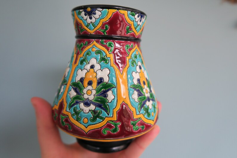 May include: A colorful ceramic vase with a geometric pattern of blue, green, yellow, white, and red flowers. The vase has a wide base and a narrow neck.