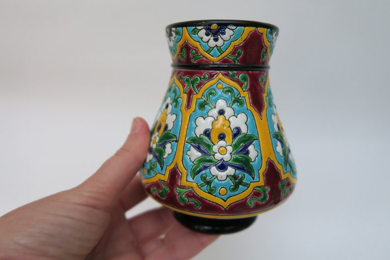 May include: A small, decorative vase with a bulbous body and a flared rim. The vase is decorated with a colorful floral pattern in shades of blue, green, yellow, and white on a burgundy background.