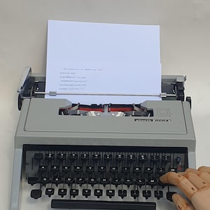 Może przedstawiać: Jasnoszara maszyna do pisania Olivetti Dora z czarną klawiaturą w układzie AZERTY i włożoną kartką białego papieru. Urządzenie posiada metalowy wózek, osłonę taśmy oraz mechaniczną konstrukcję z czarnymi klawiszami i spacją.