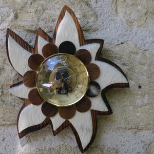May include: A decorative wall barometer in a flower shape. The barometer has a brass-colored face and is surrounded by a wooden frame with white and brown accents. The flower design is mounted on a textured stone wall.