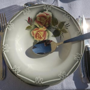 May include: A white ceramic plate with a floral design featuring yellow and pink roses. The plate is decorated with a green leafy pattern and has a white border. The plate is on a white tablecloth with a silver fork and spoon, and a black knife.