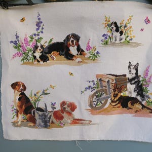 Peut inclure: Une pièce de tissu blanc avec une broderie au point de croix représentant plusieurs chiens dans un jardin fleuri. Les chiens sont de différentes races, avec une brouette en bois. La palette de couleurs comprend du marron, du noir, du blanc, du violet et du jaune.