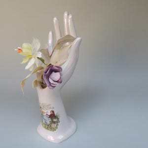 Può includere: Un vaso bianco in ceramica a forma di mano che contiene fiori artificiali. Il vaso ha un'illustrazione dettagliata alla base. I fiori includono una rosa viola, fiori bianchi e gialli e foglie beige. Il vaso è alto circa 20 cm.