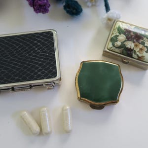 Puede incluir: Tres pastilleros decorativos en tonos plateados y dorados. Una caja tiene un patrón negro texturizado, otra tiene una tapa de esmalte verde y la tercera presenta un diseño floral. También se ven tres cápsulas.