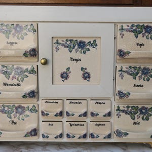 Peut inclure: Meuble à épices crème avec plusieurs tiroirs et une porte centrale. Chaque tiroir est étiqueté avec des mots français et décoré de motifs floraux bleus, verts et violets. Un bouton doré est sur la porte.