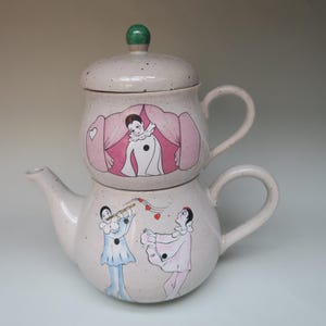 Op de afbeelding: Een tweedelige keramische theepotset met een lichtbeige gespikkelde afwerking. Het bovenste deel heeft een groene knop en een handvat. De theepot is versierd met illustraties van artiesten in roze en blauw, met een hartmotief.