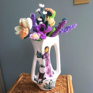Op de afbeelding: Een witte keramische kan met een handvat, met een handgeschilderde illustratie van een vrouw in een paarse jurk en een zwarte hond. De kan bevat een boeket kleurrijke, gehaakte bloemen in tinten paars, oranje, groen en wit.