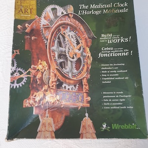 May include: A box for a 3D model kit of a medieval clock. The box features a colorful image of the assembled clock, which has a wooden frame and intricate gears. The text on the box reads "The Medieval Clock L'Horloge Médiévale" and "Build your own medieval clock. And it works!"  "Créez votre propre horloge médiévale. Elle fonctionne!"