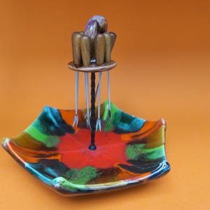 Puede incluir: Un plato de cerámica colorido con un patrón de esmalte multicolor. Un soporte de pinchos de metal con mangos de madera se encuentra en el centro del plato.