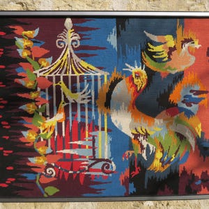 Peut inclure: Une tapisserie au point d'aiguille encadrée représentant un coq coloré et un oiseau dans une cage. La tapisserie est sur un fond sombre avec un motif floral.