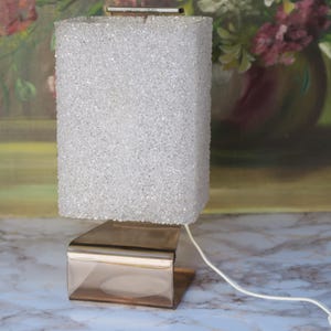 May include: A vintage table lamp with a rectangular, textured, white shade and a gold-toned metal base. The lamp has a cord with a plug. The background features a painting of flowers.