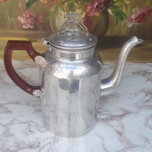 May include: A vintage aluminum coffee pot with a clear glass lid and a dark brown handle. The pot has a spout and a ridged body. The background features a painting of flowers.