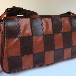 Può includere: Borsa in pelle marrone e marrone scuro con motivo a scacchiera. La borsa ha una tracolla e una chiusura con fibbia. Le cuciture sono visibili e aggiungono al design della borsa.