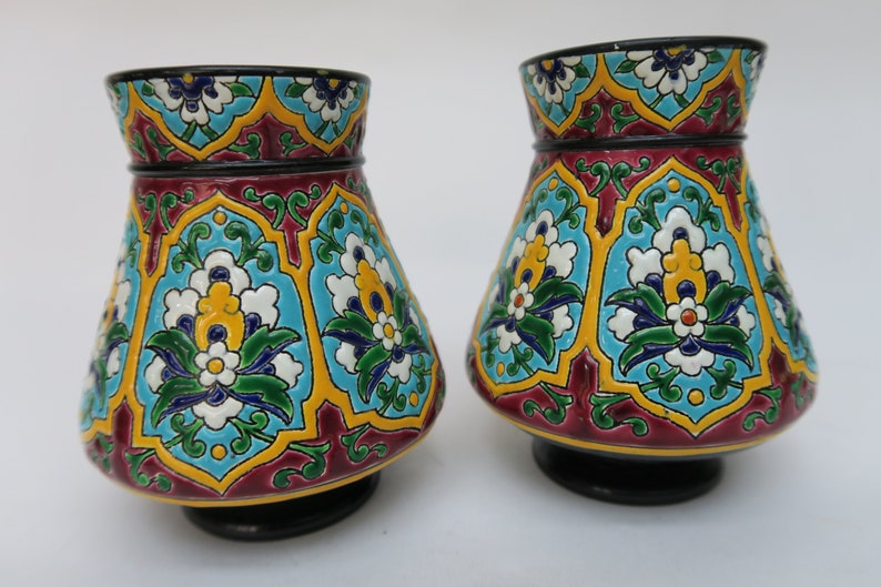May include: Two decorative ceramic vases with a colorful floral pattern. The vases are a dark red color with blue, green, yellow, and white floral designs.