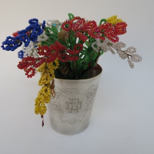 May include: A bouquet of colorful beaded flowers in a silver cup. The flowers are made of blue, red, yellow, white, and green beads.
