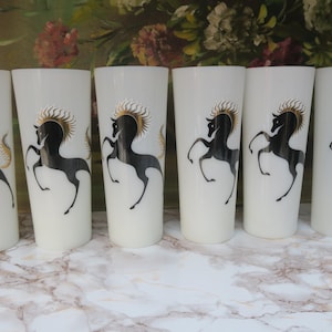 May include: Six white drinking glasses with a black horse design and gold accents. The horse is rearing up on its hind legs. The glasses are arranged in a row on a marble surface.