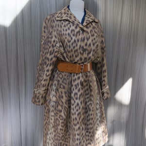 May include: A long-sleeved trench coat featuring a brown and black leopard print pattern, a pointed collar, and a wide tan leather belt with a silver-toned metal buckle cinched at the waist, displayed on a mannequin torso.