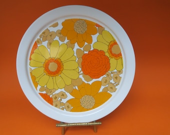 May include: A round white serving tray with a floral design. The tray features large yellow and orange flowers with brown accents on a white background. The tray is displayed on a small gold-colored stand.