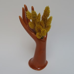 Può includere: Un vaso in ceramica a forma di mano di colore terracotta con un bouquet di fiori gialli secchi.