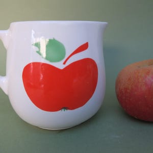 Peut inclure: Pichet en céramique blanche avec un motif de pomme rouge. La pomme a une feuille et une tige vertes. Le pichet a une anse et une forme arrondie.