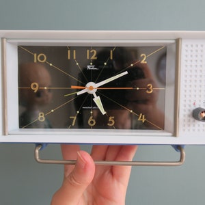 Op de afbeelding: Een witte en blauwe vintage wekker met een zwarte wijzerplaat en gouden cijfers. De klok heeft een zilveren handvat en een kleine knop aan de zijkant.