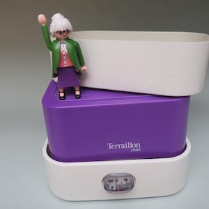 Peut inclure: Une balance de cuisine numérique blanche et violette avec une figurine Playmobil d'une femme qui salue. La balance a un dessus et un dessous blancs, un centre violet avec le texte "Terraillon 2000" et un petit cadran à l'avant.
