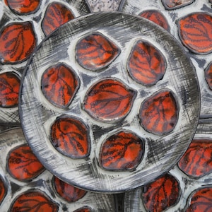 May include: Overhead view of several ceramic plates, each with multiple leaf-shaped compartments. The leaves are a vibrant red with black veining, set against a white background within a gray plate. The plates have a rustic, hand-painted appearance.