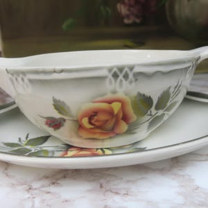 May include: A white porcelain gravy boat with a green and white floral design featuring a yellow rose. The boat is sitting on a matching saucer.
