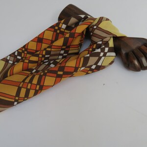 Peut inclure: Un foulard en soie à motifs géométriques de couleur marron, orange et jaune. Le foulard est noué autour d'une main en bois.