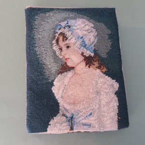 Op de afbeelding: Een naaldkunstwerk van een jong meisje dat een witte muts met blauwe bies en een witte jurk met blauw lint draagt. De achtergrond is donkerblauw.