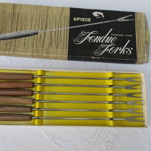 Può includere: Un set di sei forchette per fondue in una scatola. Le forchette hanno manici in legno con punte colorate e rebbi in acciaio inossidabile. La scatola è etichettata "6 pezzi Forchette per fondue Acciaio inossidabile".
