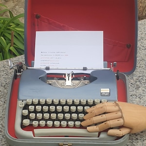 May include: A vintage blue typewriter with a white keyboard and a wooden hand prop. The typewriter is inside a grey case with a red interior. A sheet of paper with text is in the typewriter.