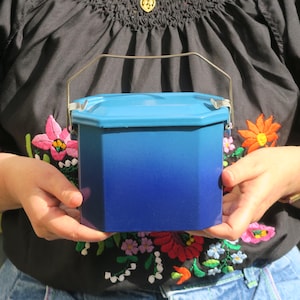 Puede incluir: Un recipiente hexagonal azul y azul claro con un asa de metal. El recipiente lo sostiene una persona que lleva una blusa negra bordada con diseños florales coloridos.