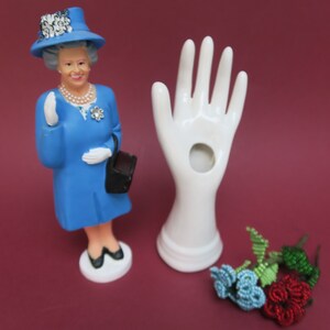 Può includere: Una statuina blu della Regina che indossa un cappello nero e tiene una borsa nera. Nell'immagine sono presenti anche un porta anelli a mano in ceramica bianca e un arrangiamento di fiori perlati.