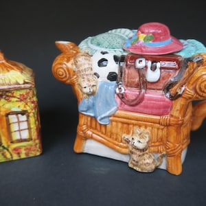 Può includere: Un bizzarro set di teiera e zuccheriera in ceramica. La teiera ha la forma di una sedia in vimini con una valigia, un cappello e altri oggetti. Lo zuccheriera assomiglia a un piccolo cottage. Entrambi i pezzi sono dipinti con colori vivaci.