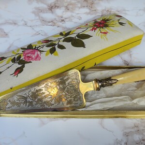 May include: A vintage cake server with a decorative floral design on the blade, presented in a gold-toned box. The server features a cream-colored handle and ornate metalwork. The box has a floral patterned lid with a yellow border.