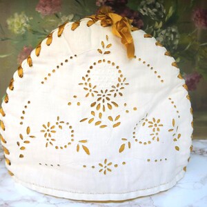 Può includere: Un copriteiera in tessuto bianco caratterizzato da un intricato ricamo a intaglio con motivo floreale. I bordi sono intrecciati con un nastro di raso color marrone dorato annodato a fiocco sulla sommità, creando una decorazione sulla fodera imbottita.